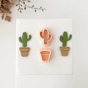 Puede incluir: Cortadores de galletas con forma de cactus y decoraciones con forma de cactus. Dos decoraciones de cactus verdes en macetas marrones y dos cortadores de galletas rosas, uno con forma de cactus y otro con forma de maceta. Sobre una superficie blanca.
