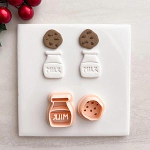 May include: A white ceramic tile with two brown cookies and two white milk bottles with the word "MILK" embossed on them. Below are two cookie cutters, one shaped like a milk bottle and the other like a cookie.