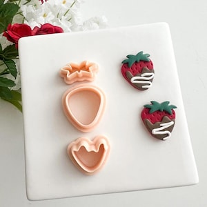 May include: A white square ceramic plate displays three pale pink cookie cutters shaped like a flower, a strawberry outline, and a strawberry base. Two chocolate-covered strawberries with green tops and white drizzle are also on the plate.