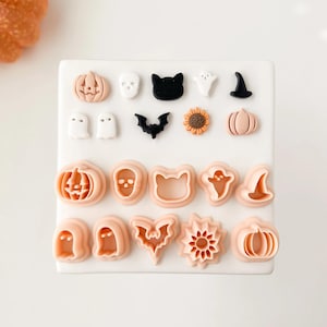 May include: A collection of Halloween-themed cookie cutters in various shapes, including pumpkins, ghosts, bats, cats, and witch hats. The cutters are in shades of peach, black, and white, arranged on a white surface.