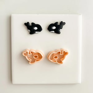 Op de afbeelding: Zwarte en witte orka-walvis oorknopjes en bijpassende koekjessnijders. De oorbellen hebben een zwarte walvis met een witte buikvlek. De koekjessnijders zijn licht perzik van kleur en hebben de vorm van een walvis.