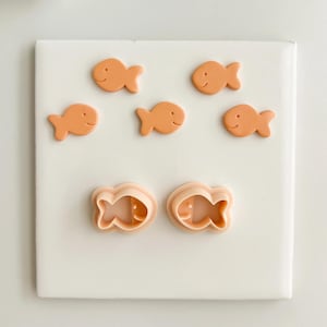 May include: A collection of orange fish-shaped cookie cutters and biscuits on a white surface. The biscuits and cutters are a light orange colour, featuring a simple fish design. The cutters are hollow, showing the shape of the fish.