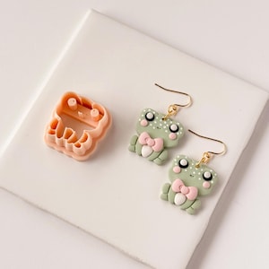 May include: A peach-coloured frog-shaped cookie cutter and two pairs of frog earrings. The earrings have gold hooks, light green bodies, pink bows, and white eyes. The earrings are a cute accessory.