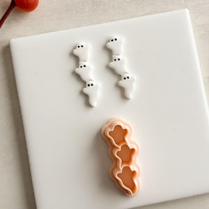 May include: White ghost-shaped decorations with black dot eyes are arranged in two vertical lines. A peach-colored ghost-shaped cookie cutter is at the bottom. The items are on a white surface. A red berry stem is in the upper left corner.