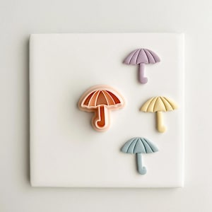 Puede incluir: Cuatro cortadores de galletas con forma de paraguas en tonos pastel melocotón, morado, amarillo y azul. Los cortadores están dispuestos sobre una superficie cuadrada blanca, ideales para hornear.