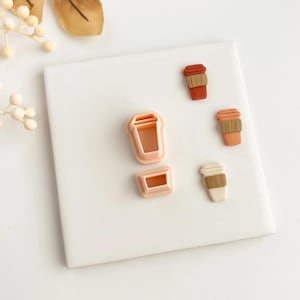 May include: A selection of coffee cup-themed items. Includes a light pink plastic cookie cutter and three miniature coffee cups in shades of brown, tan, and white, with a brown paper sleeve design. The items are arranged on a white surface.