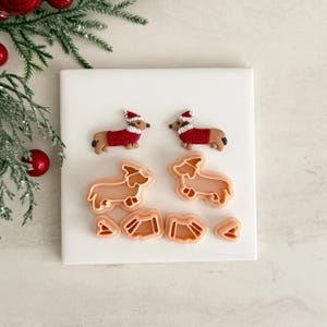 Könnte beinhalten: Eine Sammlung von Dackel-Ausstechern und dekorierten Keksen zum Thema Weihnachten. Die Kekse sind braun mit roten Pullovern und Weihnachtsmützen. Die Ausstecher sind aus hellbraunem Kunststoff. Auf einer weißen Oberfläche mit Weihnachtsdekoration.