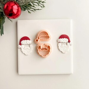 May include: A collection of Santa Claus-themed cookie cutters. The set includes cutters for Santa's hat, face, and beard. The cutters are in shades of red and peach, perfect for holiday baking. A red Christmas ornament and evergreen branches are in the background.
