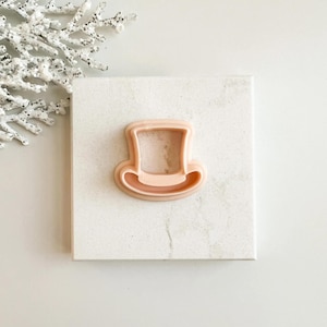 May include: A light peach-colored top hat cookie cutter. The cutter is made of plastic and sits on a white square surface. A decorative white branch is in the upper left corner.