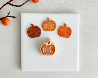 May include: Four pumpkin-shaped cookie cutters in shades of orange and brown are arranged on a white surface. A sprig with orange berries is in the upper left corner. The cutters vary in size and detail, perfect for fall baking.