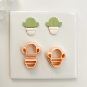 May include: Four cactus-shaped cookie cutters. Two are green with white pots, and two are peach-colored outlines of the same shape. The cutters are arranged on a white surface, perfect for baking themed treats.