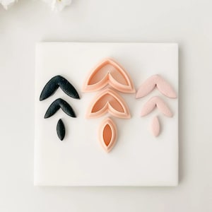 May include: A collection of leaf-shaped polymer clay cutters in black, light pink, and peach colors. The cutters are arranged on a white surface, with a few clay leaves next to them. The cutters are for making earrings or other jewelry.