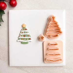 May include: A festive Christmas-themed image featuring a decorated tree, a star, and tree-shaped cookie cutters. The decorated tree is green with colorful lights and a yellow star on top. The cookie cutters are peach-colored and made of plastic.