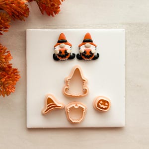 May include: Two gnome-shaped clay charms featuring orange hats and black boots, each holding a small pumpkin. Below are three light-colored plastic cookie cutters in the shapes of a witch hat, a gnome, and a small jack-o'-lantern pumpkin.