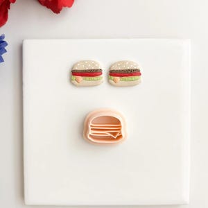 Puede incluir: Dos pendientes en miniatura con forma de hamburguesa con panecillos de sésamo, tomate rojo, lechuga y una rebanada de queso. También hay un cortador de galletas con forma de hamburguesa de color melocotón claro. Los artículos están sobre una superficie blanca.