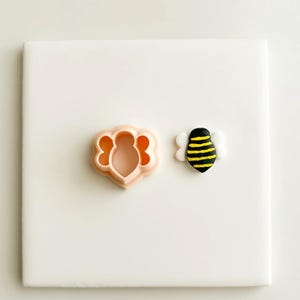 May include: A light peach-colored bee-shaped cookie cutter next to a small, hand-painted bee figurine with black and yellow stripes and white wings. The items are on a white surface.