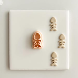 Puede incluir: Un cortador de galletas en forma de espina de pescado de color melocotón claro y tres galletas en forma de espina de pescado sobre una superficie blanca. El cortador de galletas tiene forma de esqueleto de pez. Las galletas son de color beige claro.