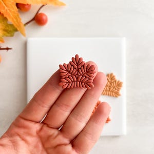 Puede incluir: Primer plano de un adorno de arcilla polimérica con forma de hoja de color terracota. El diseño detallado presenta múltiples segmentos de hojas. Adornos de hojas adicionales en colores similares son visibles sobre una superficie blanca, con follaje otoñal en el fondo.