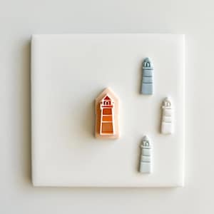 Op de afbeelding: Een verzameling koekjessnijders in de vorm van vuurtorens in verschillende kleuren, waaronder oranje, wit en blauw. De snijders zijn gerangschikt op een wit vierkant oppervlak, perfect voor het bakken van lekkernijen met kustthema's. De snijders zijn van plastic.