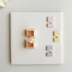 Può includere: Una collezione di oggetti geometrici dai colori pastello disposti su una superficie quadrata bianca. Le forme includono quadrati e pezzi a forma di L in tonalità di rosa, giallo e lavanda. Gli oggetti sono probabilmente decorativi o parte di un progetto artigianale.