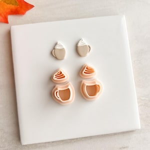 May include: Four coffee cup-shaped cookie cutters. Two are filled with white frosting, and two are empty. The cutters are light brown and peach-colored, arranged on a white surface. The image has a fall theme.