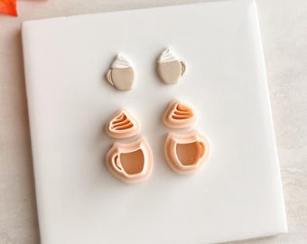 May include: Four coffee cup-shaped cookie cutters. Two are filled with white frosting, and two are empty. The cutters are light brown and peach-colored, arranged on a white surface. The image has a fall theme.