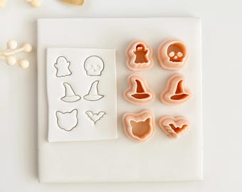 May include: A collection of Halloween-themed cookie cutters in various shapes, including a ghost, skull, witch hat, cat, and bat. The cutters are made of a light pink material and are arranged on a white surface.