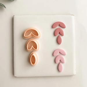 May include: A collection of polymer clay cutters in peach and pink hues. The cutters are arranged on a white surface, showcasing various leaf and petal shapes. The shapes are designed for crafting and jewelry making.