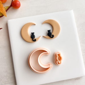 May include: A Halloween-themed image featuring two crescent moons with black cat silhouettes, and two cookie cutters. The moons are tan with black cats. The cookie cutters are pink and shaped like a crescent moon and a cat.