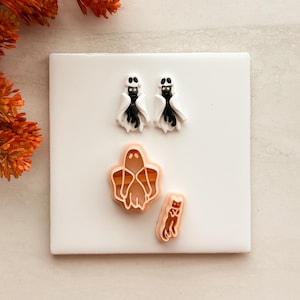 May include: Two small earrings shaped like white ghosts with black cats peeking out, featuring pink cheeks and black eyes. Below them are two peach-colored plastic cutters, one in the shape of a ghost and one in the shape of a cat, used for crafting.