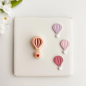 May include: Four hot air balloon-shaped cookie cutters in shades of pink and purple, arranged on a white surface. The cutters have a detailed design, with a basket and balloon shape. The background is white with a few white flowers.