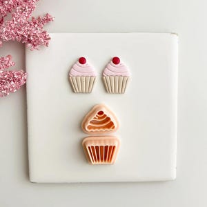 May include: Three cupcake-shaped cookie cutters. Two complete cupcakes with pink icing and a red cherry on top. The third cutter is in two pieces, showing the top and bottom of a cupcake. The cutters are peach-coloured and sit on a white surface.