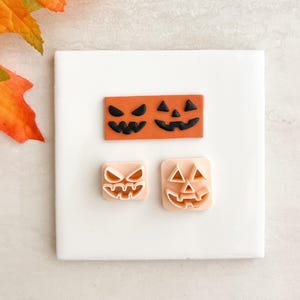Puede incluir: Tres cortadores de galletas con temática de Halloween. Dos son cuadrados y uno rectangular, todos con diseños de caras de jack-o'-lantern. Los cortadores son de color naranja y melocotón, colocados sobre una superficie blanca con hojas de otoño naranjas.