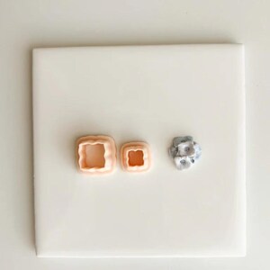May include: Three polymer clay cutters in the image. Two are square with a scalloped edge, in a peach color. The third is a small, round, multi-layered cutter in shades of gray and white. All three are on a white surface.