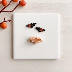 May include: A white square tile displays two black bat earrings with red flower accents and a light pink bat-shaped cookie cutter. A branch with orange berries is in the upper left corner. Halloween-themed.