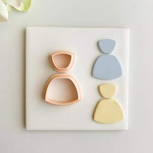 Może przedstawiać: Jasnopomarańczowa foremka do ciastek i dwa abstrakcyjne kształty w kolorze jasnoniebieskim i żółtym na białej powierzchni. Foremka do ciastek ma kształt osoby. Kształty to górna i dolna sekcja.