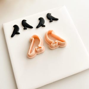 Puede incluir: Una colección de adornos negros con forma de pájaro y dos cortadores de galletas con forma de pájaro de color melocotón están dispuestos sobre una superficie blanca. Los adornos son sólidos y brillantes. Los cortadores de galletas tienen un diseño suave y redondeado.