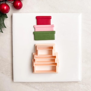 Op de afbeelding: Een verzameling kerst-thema koekjessnijders. De snijders hebben de vorm van gestapelde geschenkdozen in rood, roze en groen. Er is ook een licht perzikkleurige snijder aanwezig. Rode bessen en groene bladeren bevinden zich in de linkerbovenhoek.