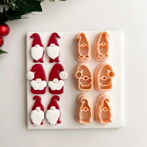 May include: A collection of gnome-shaped cookie cutters and decorated cookies. The cookies feature red hats and white beards. The cutters are in a light peach color. The items are arranged on a white surface, with holiday greenery in the background.