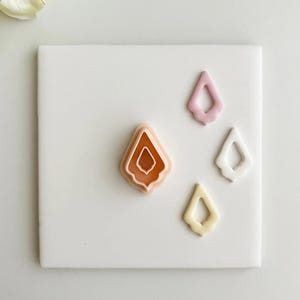Könnte beinhalten: Vier tropfenförmige Ohrringkomponenten aus Polymer Clay in Pfirsich-, Rosa-, Weiß- und Gelbtönen. Die Komponenten sind auf einer weißen quadratischen Fliese angeordnet. Die größte Komponente hat ein geschichtetes Design.