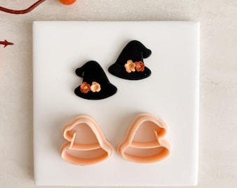 Op de afbeelding: Twee zwarte heksenhoeden met oranje en gele bloemaccenten en twee lichtroze heksenhoedvormige koekjessnijders op een wit oppervlak. Voor Halloween- of herfstgebak.