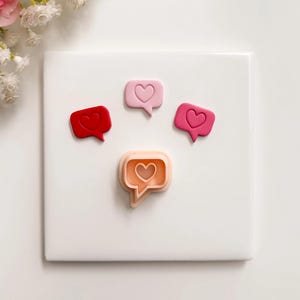 以下が含まれることがあります： 赤、ピンク、ピーチの色合いのハートアイコンが付いた、吹き出し型のオブジェクトが4つ、白い正方形に配置されています。これらは、バレンタインデーやソーシャルメディアをテーマにした装飾品または工芸品である可能性があります。