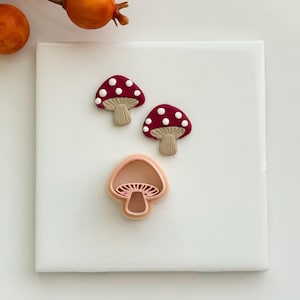 May include: Two small mushroom-shaped charms with deep red caps featuring white polka dots and tan ribbed stems, accompanied by a single light peach-colored plastic mushroom-shaped cookie cutter with a detailed interior gill design.