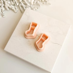 Puede incluir: Dos cortadores de galletas de color melocotón claro con forma de calcetines navideños. Los cortadores son de plástico y están colocados sobre una superficie de mármol blanco. Una rama decorativa blanca y brillante está en el fondo.