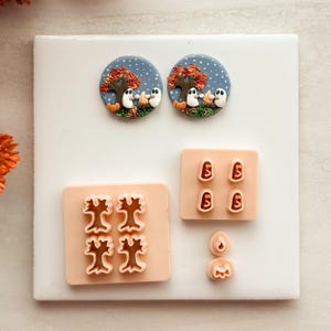 May include: Two circular clay charms featuring a night scene with ghosts, a campfire, and autumn trees. Below are peach-coloured plastic cutters for these shapes, including four tree cutters, four ghost cutters, and two small flame-shaped cutters.