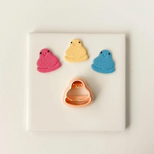 May include: A collection of colorful chick-shaped cookie cutters and treats. The image features a pink, yellow, and blue chick-shaped treats, along with a peach-colored cookie cutter. The treats have small black dot eyes.