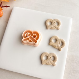 Puede incluir: Un cortador de galletas de pretzel de color beige claro y tres pretzels horneados. El cortador de galletas es de color melocotón claro. Los pretzels están sobre una superficie blanca con follaje otoñal en el fondo.