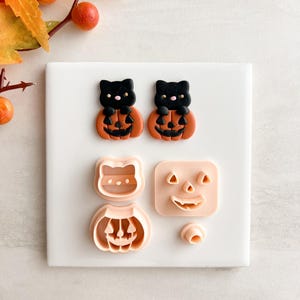 May include: A selection of Halloween-themed cookie cutters and decorations. Two black cat figures sit atop orange pumpkins. Additional cutters include a cat face, a pumpkin, and a jack-o'-lantern face. A small, round piece is also included.