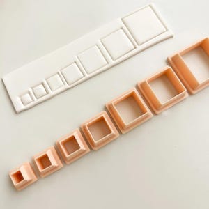 Puede incluir: Un conjunto de cortadores de galletas cuadrados de varios tamaños, dispuestos en fila. Los cortadores son de color melocotón, y también hay un molde rectangular blanco con hendiduras cuadradas. Los artículos están sobre una superficie blanca.