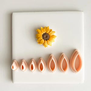 May include: A yellow and brown sunflower and a set of seven pink petal-shaped cookie cutters arranged on a white surface. The sunflower is in the center, and the cutters are below it.